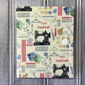 May include: A rectangular fabric pouch with a cream background featuring a repeating pattern of sewing machines, spools of thread, hangers, scissors, safety pins, and the text 100% Handmade. The pouch is secured with a single white plastic snap button.