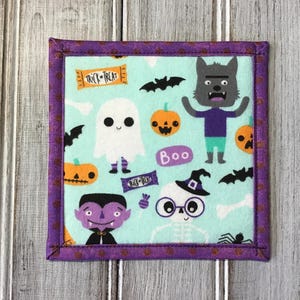 May include: A square fabric coaster with a light mint green background featuring a Halloween pattern of ghosts, pumpkins, bats, a werewolf, a vampire, and a skeleton. The text "TRICK OR TREAT" and "BOO" is printed on the fabric. It has a purple border.