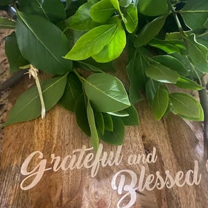 May include: A wooden cutting board with the words "Grateful and Blessed" in a cursive font. Fresh green leaves are arranged on top of the board, creating a natural and inviting aesthetic. The board's wood grain adds texture.