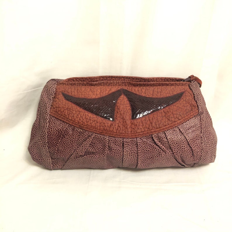 May include: A brown leather clutch purse with a textured finish. The front flap has a contrasting brown leather design with a black leather center.