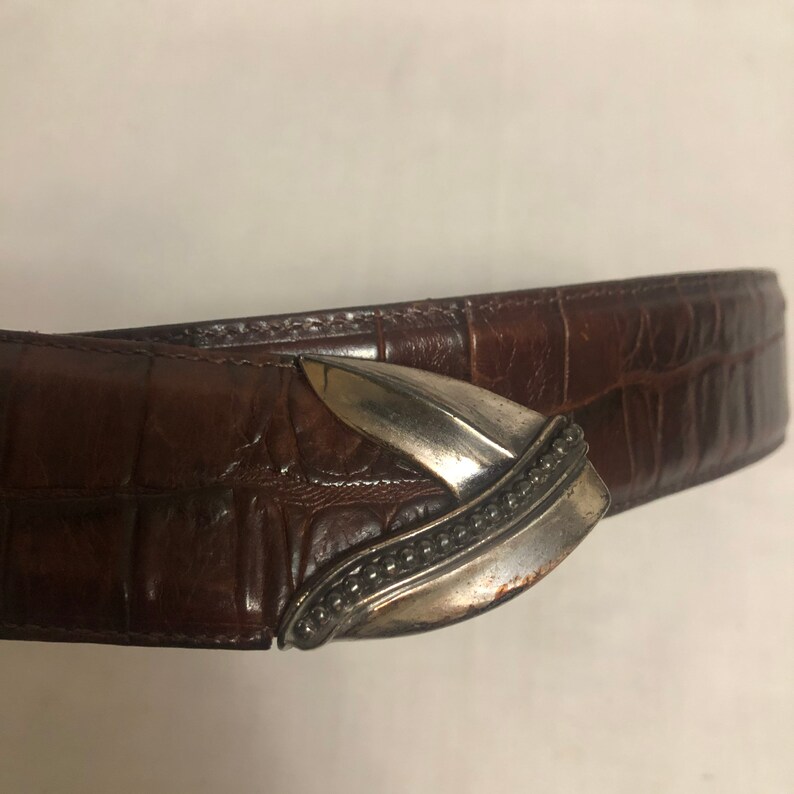 以下が含まれることがあります： 先のとがった歯の形をした銀色のバックルが付いた茶色の革ベルト。バックルにはビーズの装飾的な縁取りが施されています。