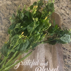 May include: A wooden cutting board with the text "Grateful and Blessed" inscribed on it. A bunch of green leafy stems with yellow flowers are arranged on the board.