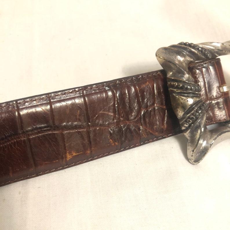 以下が含まれることがあります： 茶色の革製のベルトに銀色のバックルが付いている。バックルはねじれたデザインで、装飾的なパターンが施されている。