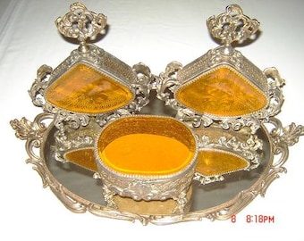 May include: A silver-tone vanity set with three compartments. The compartments are shaped like triangles and have a gold-tone finish. The set is decorated with ornate details and has a mirrored base.