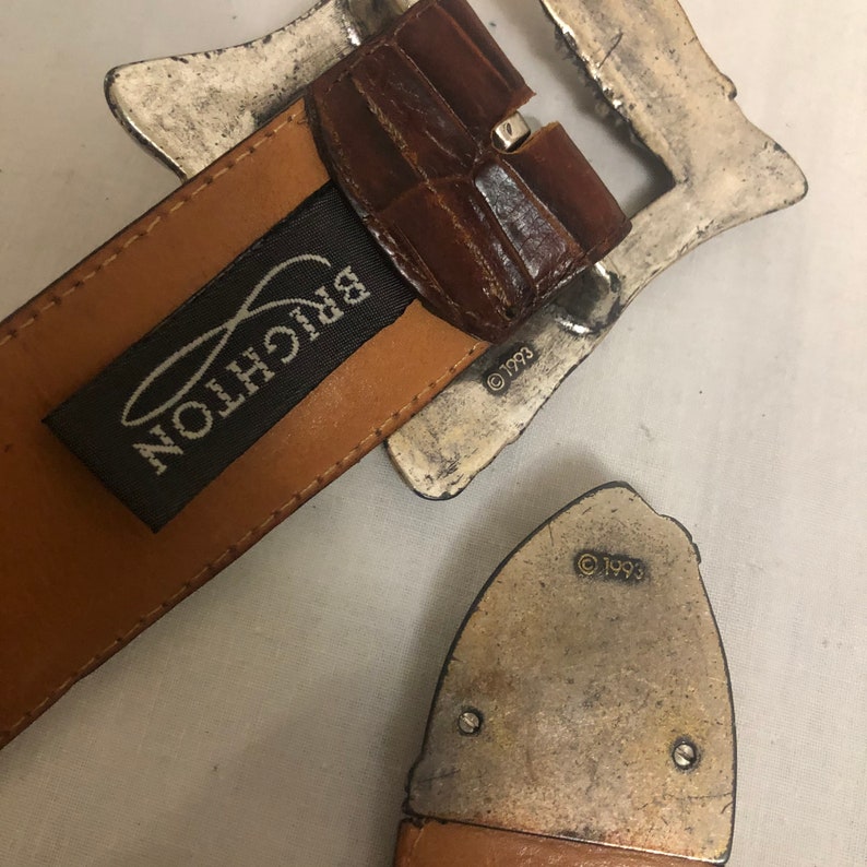 以下が含まれることがあります： 茶色の革製のベルトに銀色のバックルが付いている。バックルは装飾的なデザインで、ベルトには「BRIGHTON」という文字が見える。バックルには著作権マークと1993年が刻印されている。