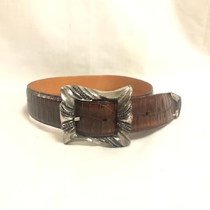 以下が含まれることがあります： 銀色のバックルが付いた茶色の革ベルト。バックルは渦巻き模様の装飾的なデザインです。