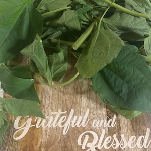 May include: A wooden cutting board with a light brown grain pattern features the white script text Grateful and Blessed. A cluster of fresh green leaves with visible stems and veins is arranged across the top portion of the wooden surface.