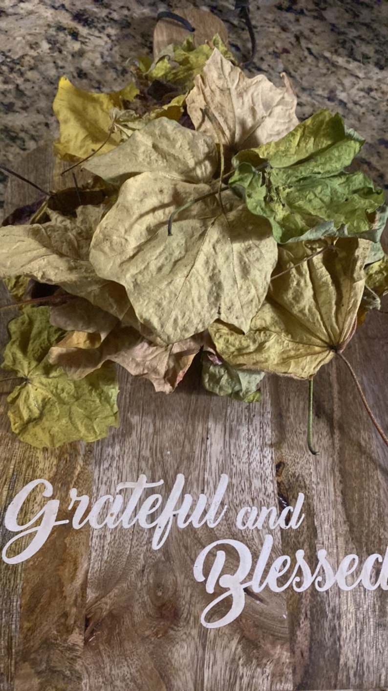 Op de afbeelding: Een houten bord met de tekst "Grateful and Blessed" in witte letters. Een stapel gedroogde bladeren in verschillende tinten geel, bruin en groen liggen op het bord.