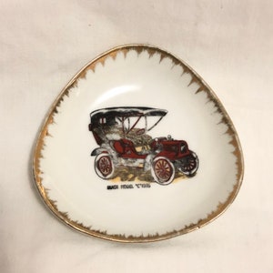 May include: A white porcelain trinket dish with a gold rim. The dish features a vintage red car with a black top and the text "BUICK MODEL "C" 1905".