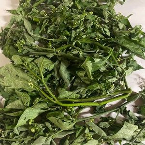 May include: A pile of fresh green leafy plants with small, round, green berries. The leaves are a deep green color, and the stems are a lighter green. The berries are clustered together. The plants are on a white surface.