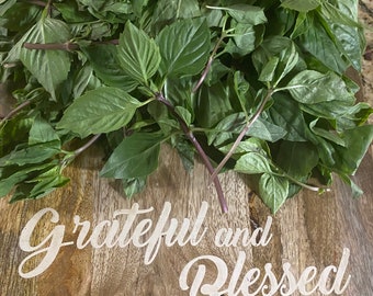 May include: A wooden cutting board with a pile of fresh green basil leaves and the text "Grateful and Blessed" written in white.