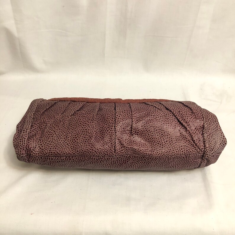 May include: A dark purple clutch purse with a textured, pebbled leather exterior. The purse has a pleated design and a slightly rounded shape.