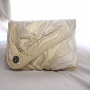 May include: A white leather clutch bag with a decorative, abstract design. The bag has a silver button closure.