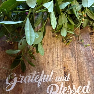 May include: A wooden cutting board with the words "Grateful and Blessed" in white script. Green leafy branches are arranged across the top of the board. The board has a natural wood grain.