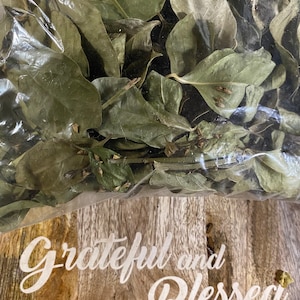 May include: A clear plastic bag filled with dried green leaves. The bag is sitting on a wooden surface with the text "Grateful and Blessed" written in white.