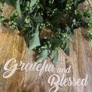 May include: A wooden cutting board with the words "Grateful and Blessed" in white script. Green leaves and berries are arranged on the board, with some berries appearing black and others green.