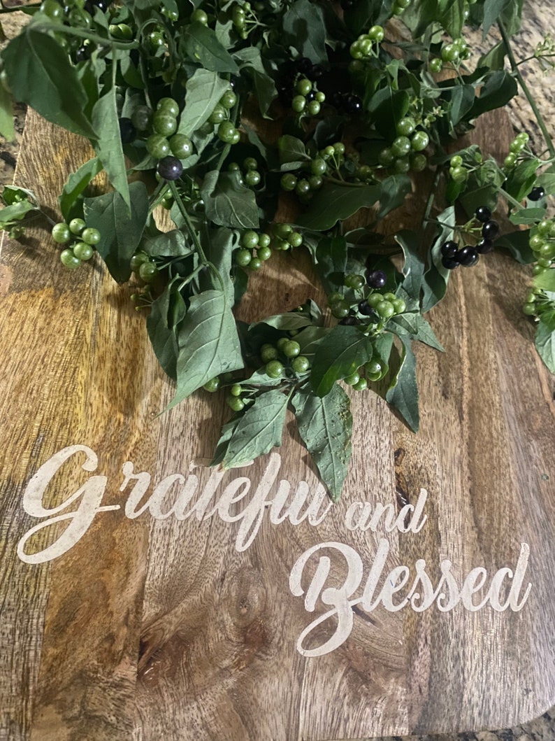 May include: A wooden cutting board with the words "Grateful and Blessed" in white script. Green leaves and berries are arranged on the board, with some berries appearing black and others green.