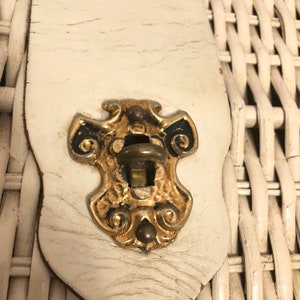 Puede incluir: Primer plano de una cesta de mimbre blanca con un cierre de metal dorado y negro. El cierre tiene un dise&ntilde;o decorativo con espirales y un peque&ntilde;o pomo.