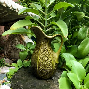 May include: A green ceramic pitcher with an embossed floral design. The pitcher is sitting on a rock with green plants in the background.