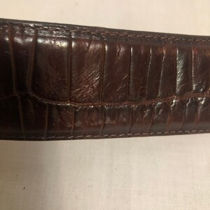 以下が含まれることがあります： クロコダイル型押し模様の茶色の革ベルトのクローズアップ。ベルトはステッチされた縁を持っています。