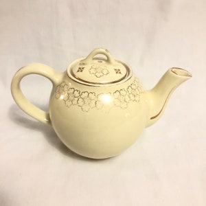 May include: A cream-colored teapot with a gold floral design and a gold trim around the lid. The teapot has a round body and a curved spout.
