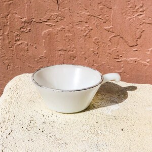May include: A white ceramic bowl with a handle. The bowl has a slightly flared rim and a gray rim around the top.