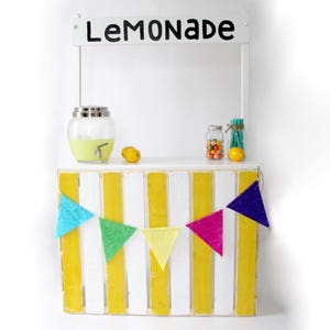 May include: A white wooden lemonade stand with a yellow and white striped front. The stand has a sign that says 