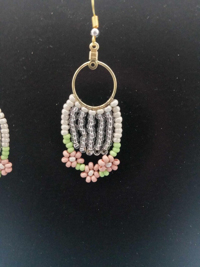 May include: A pair of dangle earrings with a gold hoop and a beaded fringe. The fringe is made of white, clear, and green beads. The bottom of the fringe is decorated with pink flowers.