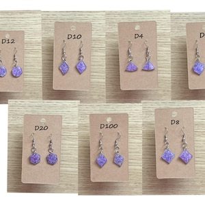 May include: A collection of dice-shaped earrings in various styles, including D12, D10, D4, D6, D20, D100, and D8. Each earring features a purple die with white numbering, suspended from a silver hook. The earrings are displayed on small, brown cardboard tags.