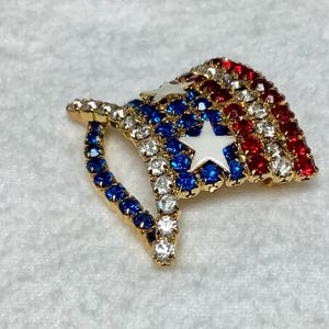 May include: A gold-tone brooch in the shape of an American flag. The flag is made of red, white, and blue rhinestones. The brooch is a patriotic accessory.