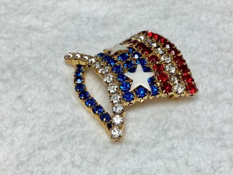 May include: A gold-tone brooch in the shape of an American flag. The flag is made of red, white, and blue rhinestones. The brooch is a patriotic accessory.
