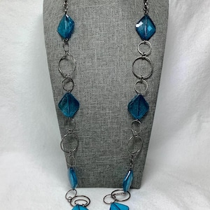 May include: A long necklace featuring a dark metal chain interspersed with translucent blue diamond-shaped beads and circular metal rings. The design alternates between the geometric blue beads and the dark metallic circles, creating a layered look.