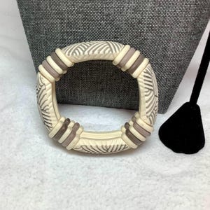 May include: Ivory-colored bangle bracelet with a repeating pattern of gray lines and brown accents. The bracelet has a circular shape and is made of a solid material. The bracelet is a fashion accessory.