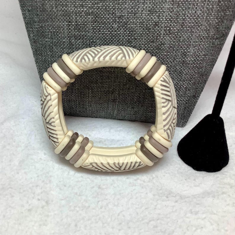 May include: Ivory-colored bangle bracelet with a repeating pattern of gray lines and brown accents. The bracelet has a circular shape and is made of a solid material. The bracelet is a fashion accessory.