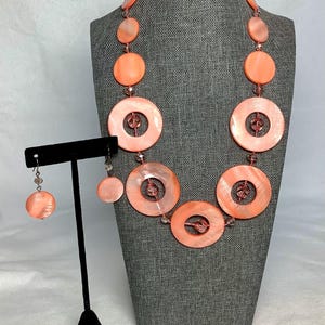 以下が含まれることがあります： サンゴ色のネックレスとイヤリングのセット。ネックレスは、大きな円形のオープンセンタービーズと、それらに挟まれた小さな丸いビーズが特徴です。イヤリングは、1つの小さな円形ビーズが付いています。ジュエリーはグレーのマネキンに展示されています。