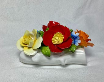 Könnte beinhalten: Eine weiße Porzellan-Blumenarrangements mit einem roten, gelben und orangefarbenen Blumenstrauß. Die Blumen sind auf einem weißen Porzellanast angeordnet.