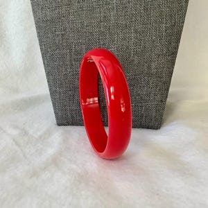 May include: A vibrant red bangle bracelet. The smooth, glossy surface reflects light, highlighting its circular form. The bracelet is a bold accessory, perfect for adding a pop of color to any outfit. The bangle is approximately 3 inches in diameter.