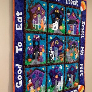 K&ouml;nnte beinhalten: Ein Halloween-Quilt mit neun quadratischen Feldern, die Spukh&auml;user, Geister und Trick-or-Treat-Kinder darstellen. Der Quilt enth&auml;lt die S&auml;tze "Trick or Treat", "Good To Eat", "Smell My Feet" und "Give Me Something". Der Rand ist blau mit orangefarbenen Akzenten und Zuckermais.