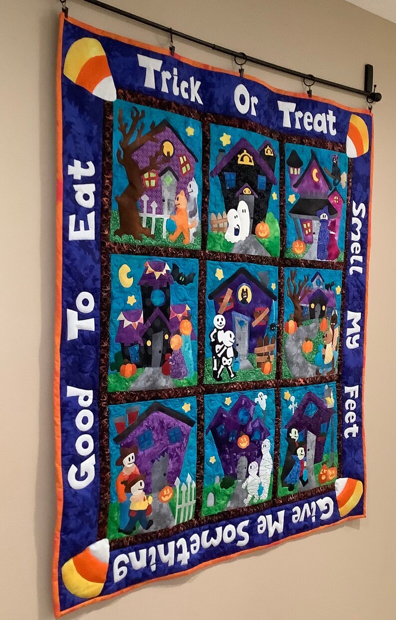 K&ouml;nnte beinhalten: Ein Halloween-Quilt mit neun quadratischen Feldern, die Spukh&auml;user, Geister und Trick-or-Treat-Kinder darstellen. Der Quilt enth&auml;lt die S&auml;tze "Trick or Treat", "Good To Eat", "Smell My Feet" und "Give Me Something". Der Rand ist blau mit orangefarbenen Akzenten und Zuckermais.