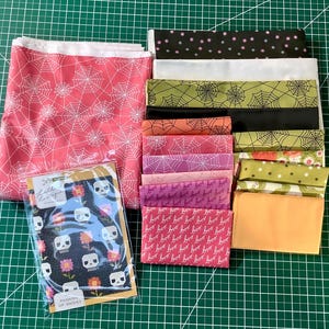 May include: A collection of Halloween-themed fabric bundles. The fabrics feature spiderwebs, skulls, and the word "boo" in various colors, including pink, black, orange, and green. A small package with skull and flower print is also included.
