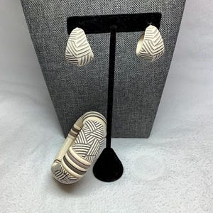 May include: A set of cream-colored earrings and a matching bracelet with a geometric design. The earrings are teardrop-shaped, and the bracelet is a wide, rounded bangle. The jewelry features a pattern of dark gray lines.