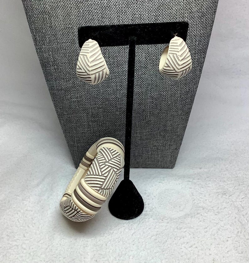 May include: A set of cream-colored earrings and a matching bracelet with a geometric design. The earrings are teardrop-shaped, and the bracelet is a wide, rounded bangle. The jewelry features a pattern of dark gray lines.