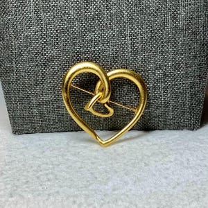 May include: A gold-toned heart-shaped brooch with a smaller heart design inside. The brooch is made of metal and has a pin on the back.