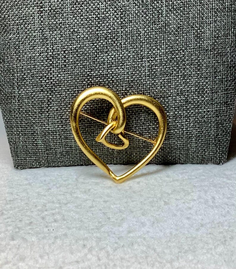 May include: A gold-toned heart-shaped brooch with a smaller heart design inside. The brooch is made of metal and has a pin on the back.