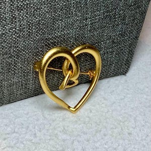 May include: A gold-toned heart-shaped brooch with an intricate design. The brooch is made of metal and has a shiny finish.