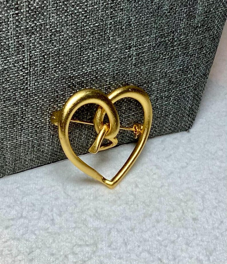 May include: A gold-toned heart-shaped brooch with an intricate design. The brooch is made of metal and has a shiny finish.