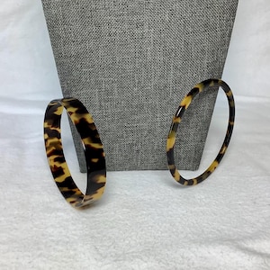 May include: Two circular bangle bracelets featuring a classic brown and black tortoiseshell pattern. One bangle is wider with a thicker profile, while the other is a thinner, more delicate band. Both bracelets have a smooth, polished finish.