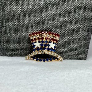 May include: A gold-tone pin shaped like a top hat with red, white, and blue rhinestones. The hat has two white stars on the front.