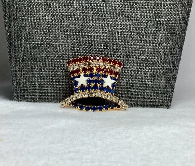 May include: A gold-tone pin shaped like a top hat with red, white, and blue rhinestones. The hat has two white stars on the front.