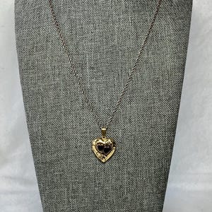 May include: A gold-colored heart-shaped locket necklace. The locket hangs from a delicate chain and features a detailed design. The necklace is displayed on a gray mannequin.